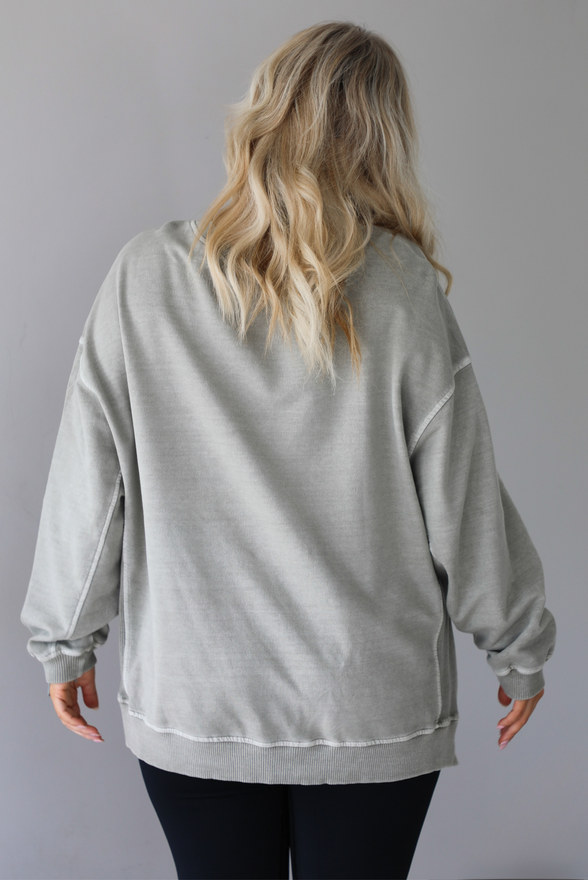 Woman wearing a tan oversized sweater with black leggings that have a slightly flared hem with split hem details in the front. The photo shows a closeup of the back of the sweater and has a white background. 