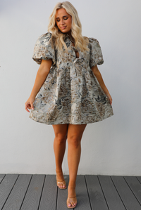 Woman model wearing a champagne and multi-colored floral pattern throughout. The dress has puffed sleeves, a v-neckline with tie details, and a baby doll cut that stops mid thigh on the 5'7" mode. The photo is taken outdoors against a white wall. 