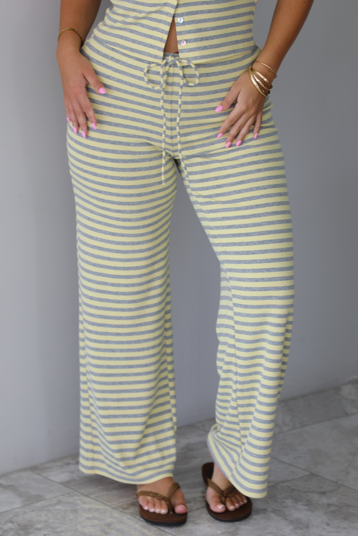 A woman wearing a two piece set consisting of a collared, button-down, short sleeve top and high waisted, drawstring, wide leg pants, standing indoors against a white wall with white marble flooring. It shows a closeup of the front of the pants.