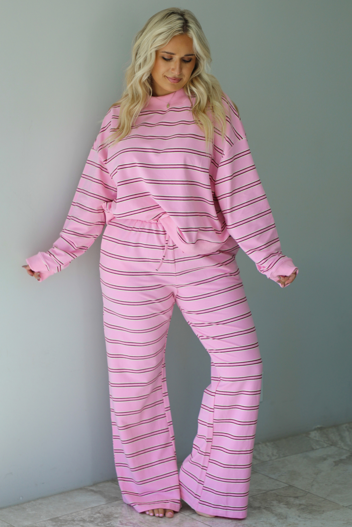 A woman wearing a pink two piece sweat set featuring a maroon and white striped design on a sweatshirt and high waisted, wide leg pants, standing indoors against a white wall with white marble flooring. 