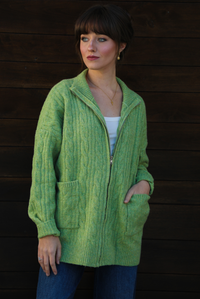 Woman model wearing a green cable knit zip-up cardigan with dark denim jeans outdoors against a dark brown wooden door. The model is wearing a Small. 