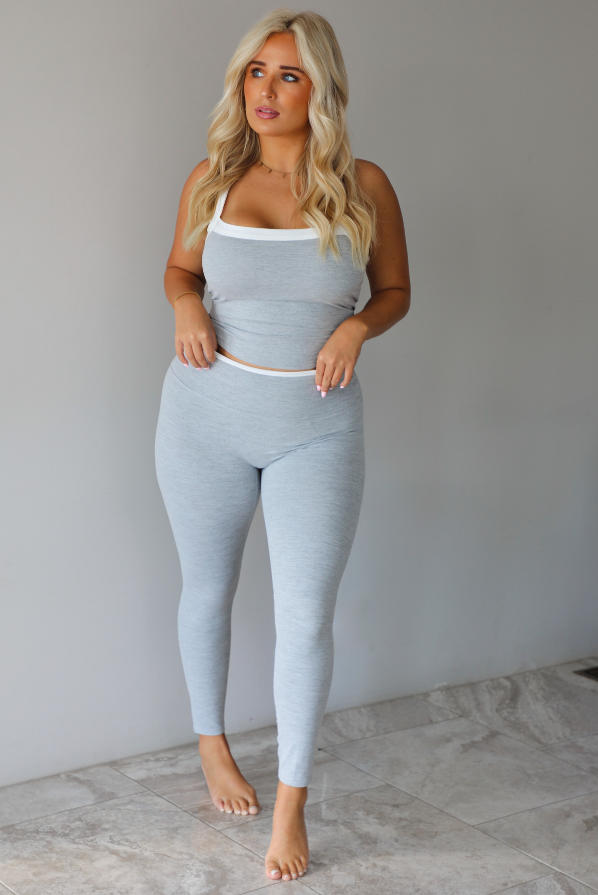 A woman modeling a light gray two-piece loungewear set consisting of a cropped tank top and matching leggings, standing indoors against a white wall with white marble flooring. 