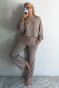 A woman wearing a mocha and white striped matching two piece set featuring a cropped, flowy. long sleeved top paired with high waisted wide leg pants, standing indoors against a white wall with white marble flooring, taking a mirror selfie. 