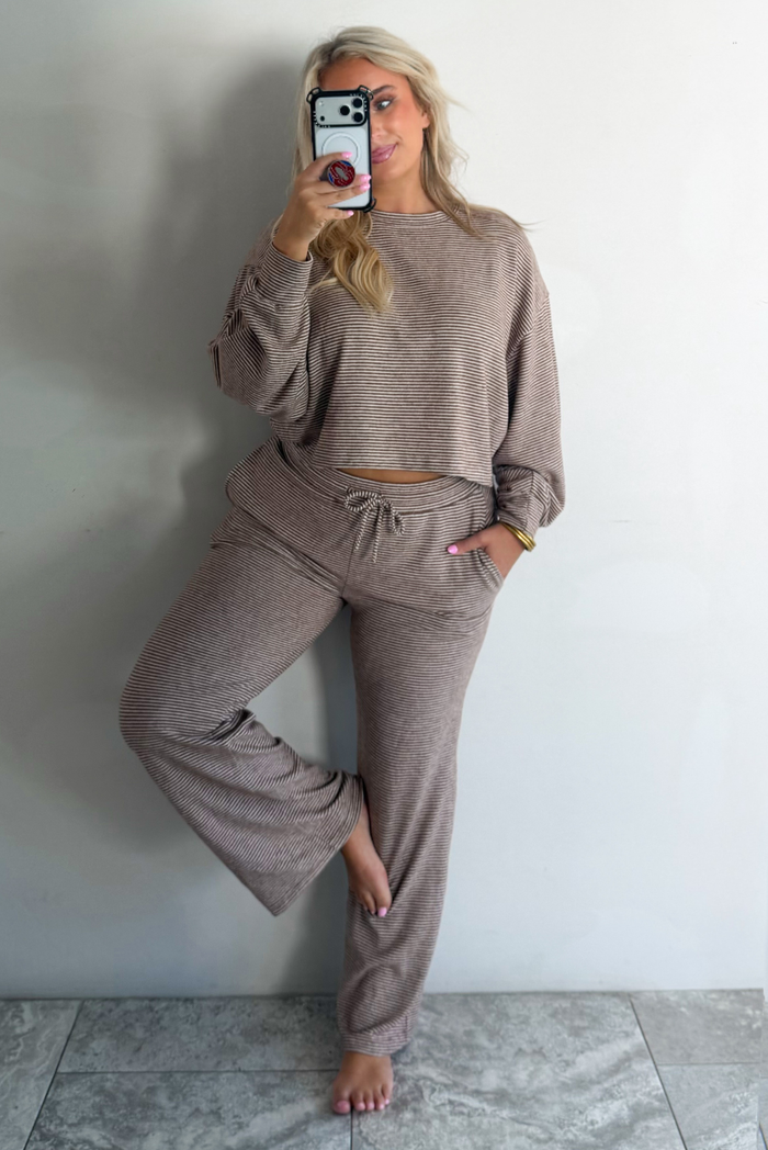 A woman wearing a mocha and white striped matching two piece set featuring a cropped, flowy. long sleeved top paired with high waisted wide leg pants, standing indoors against a white wall with white marble flooring, taking a mirror selfie. 