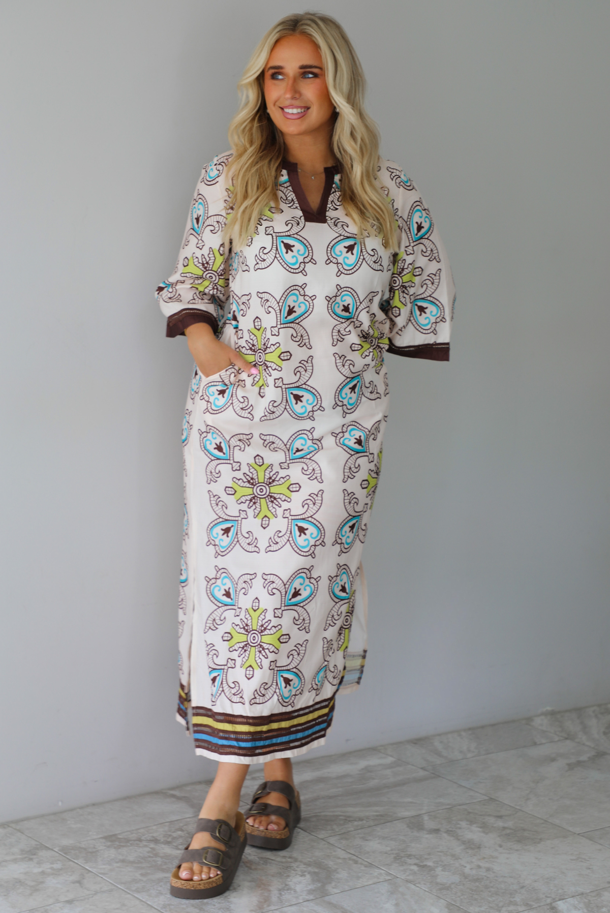 A woman modeling a long kaftan dress with a colorful paisley pattern on a cream background, featuring brown trim and sandals, standing indoors against a white wall with white marble flooring. 