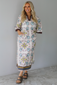 A woman modeling a long kaftan dress with a colorful paisley pattern on a cream background, featuring brown trim and sandals, standing indoors against a white wall with white marble flooring. 