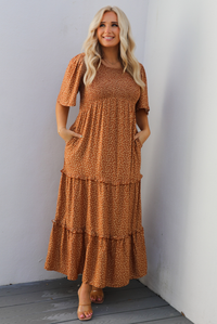A woman model outdoors wearing a rust dress with a cream floral print pattern throughout. It has flowy short sleeves, a smocked bodice, and a flowy maxi length skirt with functional pockets on either side. 