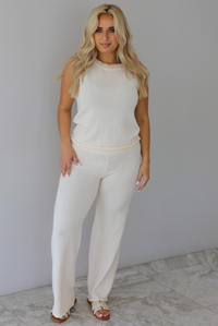 Woman wearing a cream two piece set featuring a high-neck sleeveless top and matching mid-rise pants indoors against a white wall with white marble flooring. 