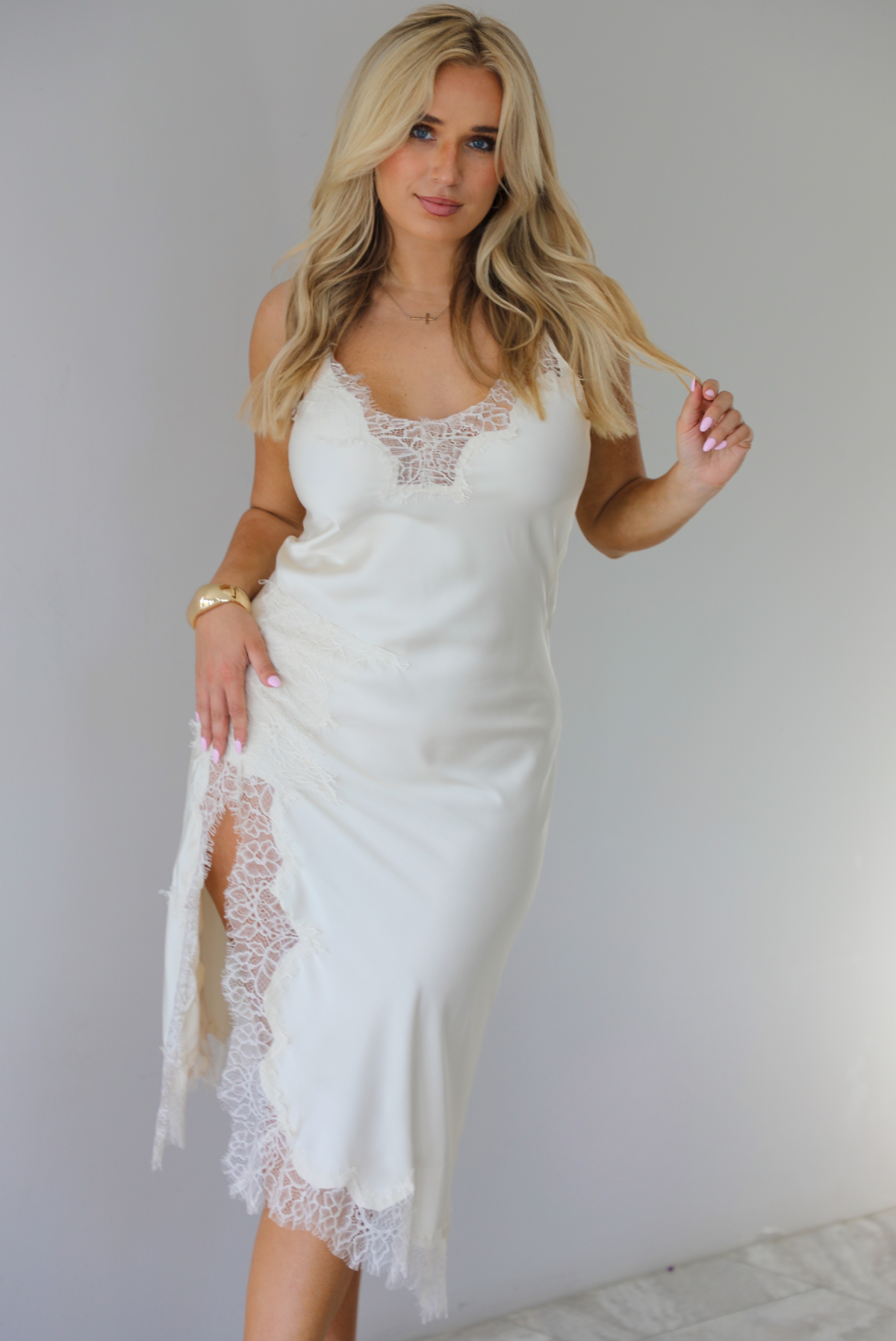 Woman wearing an off white satin midi length dress that has spaghetti straps, lace detailing at the v-neckline, and lace detailing at the hem and split hem detail on the right side. The photo is taken indoors against a white wall with white marble flooring. 