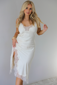 Woman wearing an off white satin midi length dress that has spaghetti straps, lace detailing at the v-neckline, and lace detailing at the hem and split hem detail on the right side. The photo is taken indoors against a white wall with white marble flooring. 