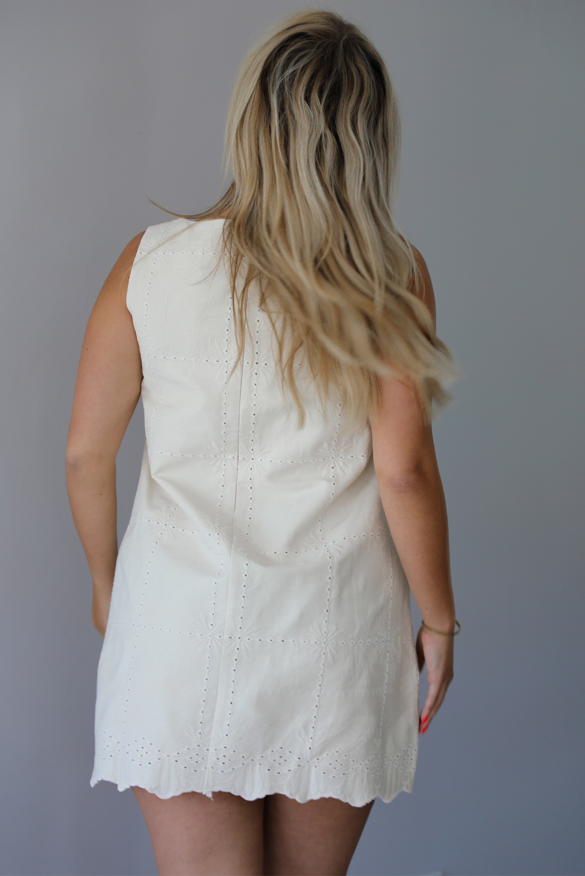 Woman wearing a sand colored mini dress with a square neckline, eyelet details throughout, and a scalloped hem indoors against a white wall. The photo shows the back of the dress, which has a zipper closure. 
