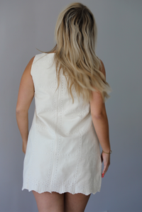 Woman wearing a sand colored mini dress with a square neckline, eyelet details throughout, and a scalloped hem indoors against a white wall. The photo shows the back of the dress, which has a zipper closure. 