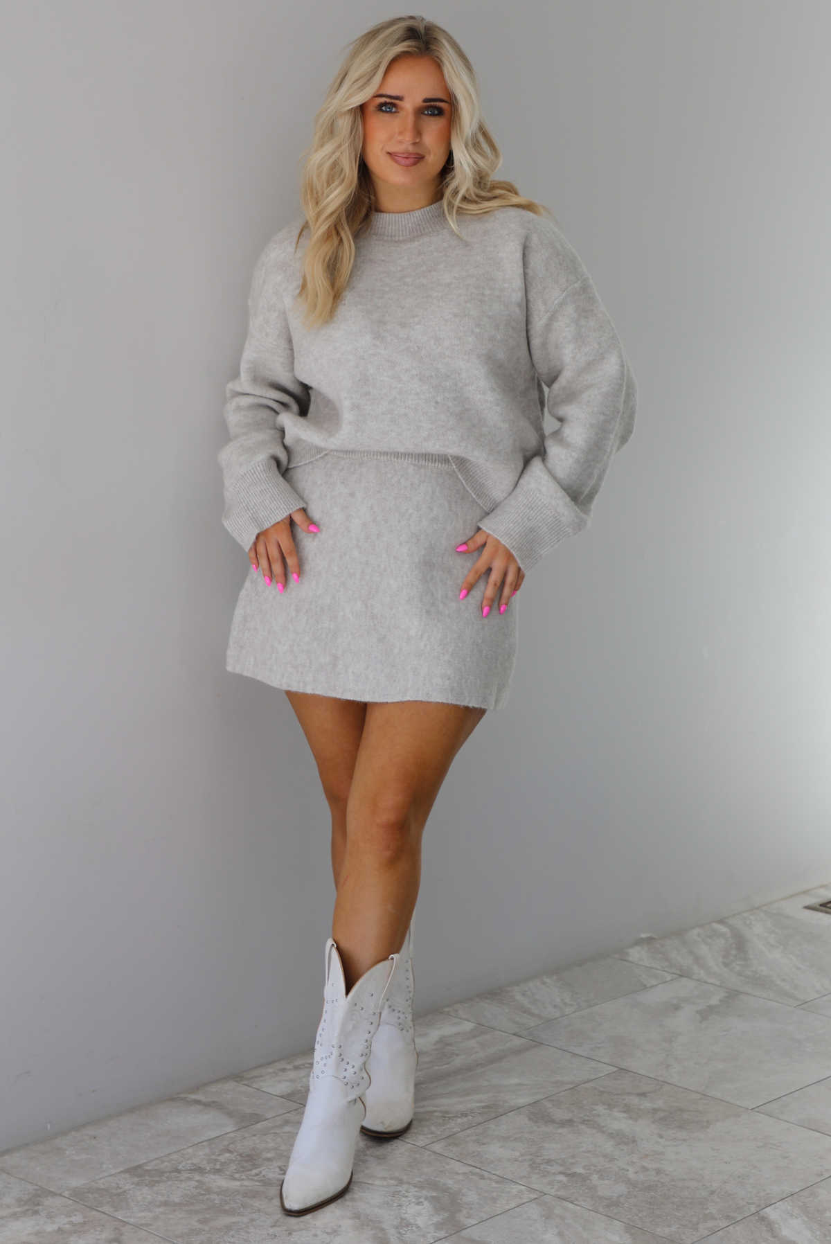 Woman wearing a grey two piece sweater set featuring a high neck sweater and a skirt. The photo is taken indoors against a white wall with white marble flooring. 