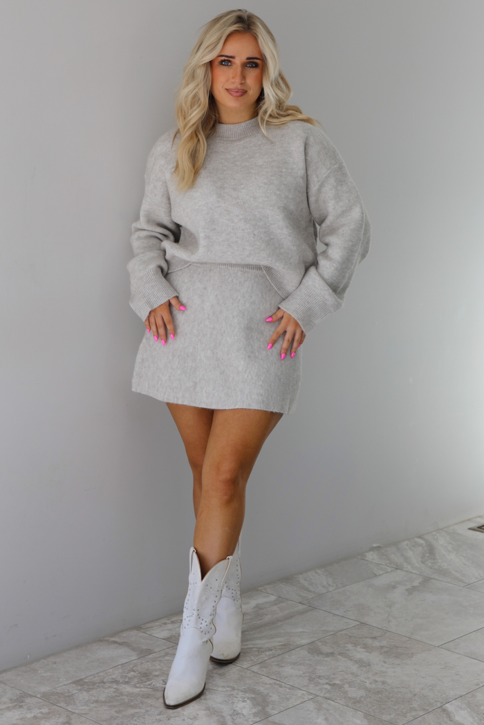 Woman wearing a grey two piece sweater set featuring a high neck sweater and a skirt. The photo is taken indoors against a white wall with white marble flooring. 