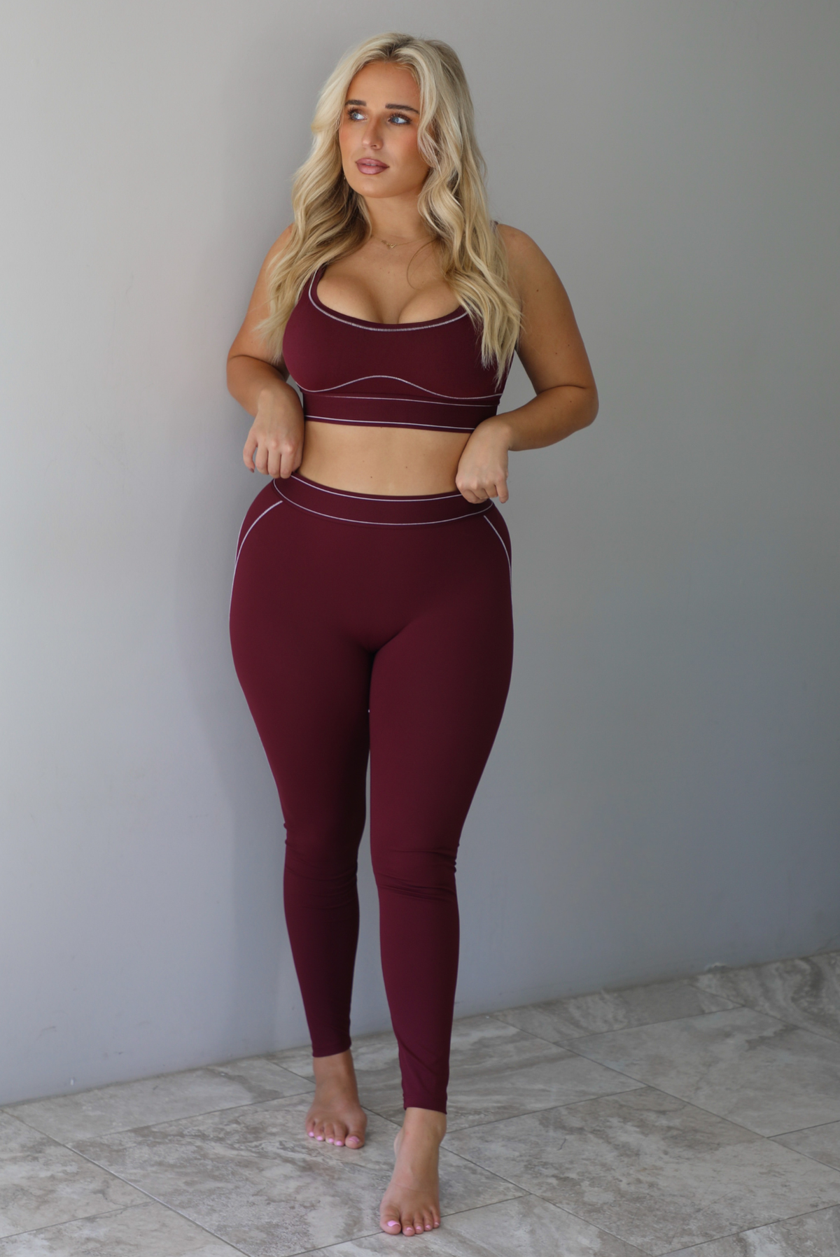 A woman wearing a maroon matching two piece set featuring a sports bra and high waisted leggings with white stitching details, standing indoors against a white wall with white marble flooring. 
