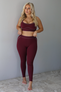 A woman wearing a maroon matching two piece set featuring a sports bra and high waisted leggings with white stitching details, standing indoors against a white wall with white marble flooring. 