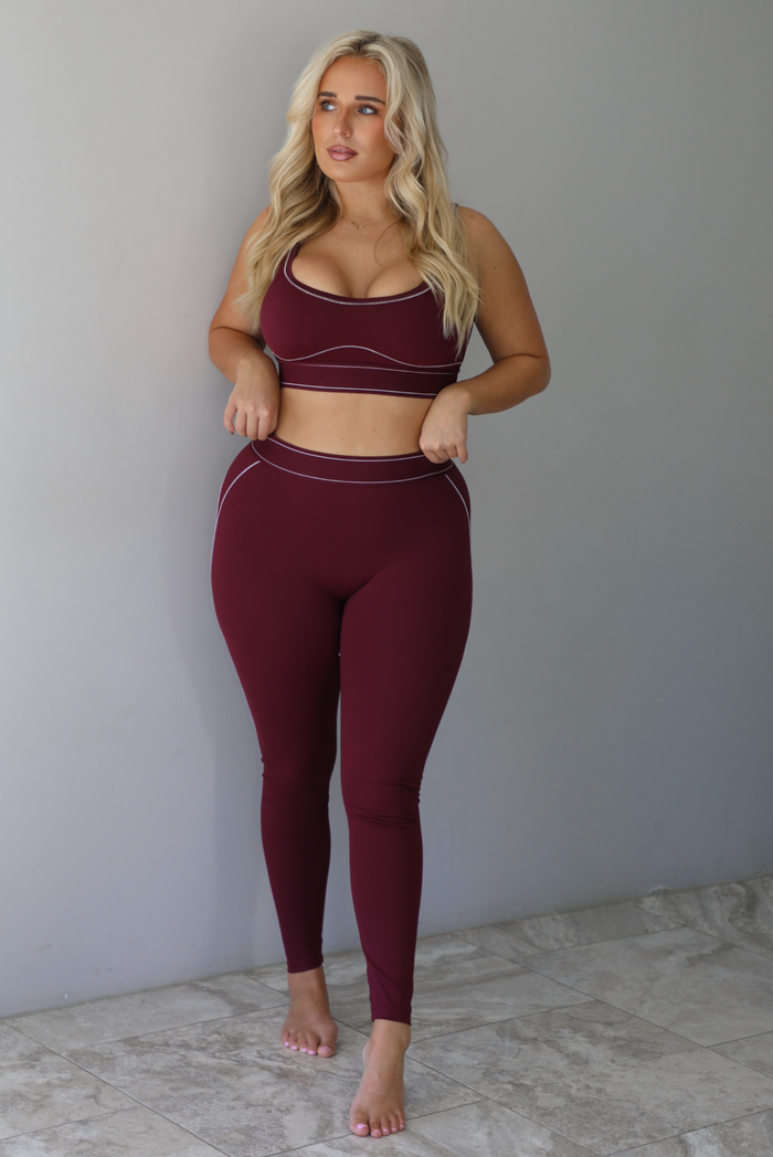 A woman wearing a maroon matching two piece set featuring a sports bra and high waisted leggings with white stitching details, standing indoors against a white wall with white marble flooring. 