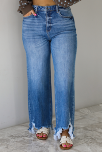 Woman wearing a medium wash denim that are high waisted, have functional front and back pockets, functional button and zipper closures, and have a raw, distressed hem. The photo is taken indoors against a white wall with white marble flooring.