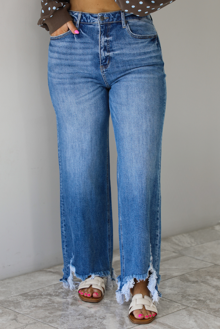 Woman wearing a medium wash denim that are high waisted, have functional front and back pockets, functional button and zipper closures, and have a raw, distressed hem. The photo is taken indoors against a white wall with white marble flooring.