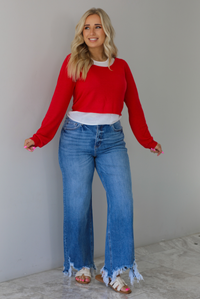 Woman wearing a red and white layered appearance top with dark wash denim jeans. The photo is taken indoors against a white wall with white marble flooring. 