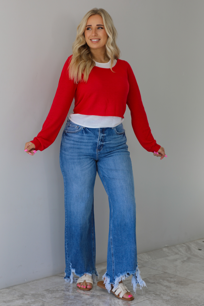 Woman wearing a red and white layered appearance top with dark wash denim jeans. The photo is taken indoors against a white wall with white marble flooring. 