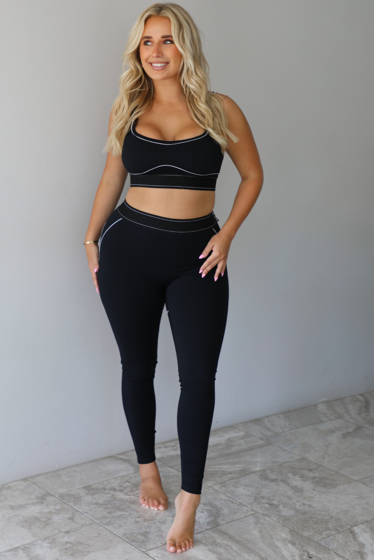 A woman modeling a matching black activewear set consisting of a sports bra and high-waisted leggings with thin white contrast stitching, standing indoors against a white wall with white marble flooring. 