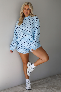 A woman wearing a blue two piece set with a navy polka dot pattern throughout, featuring a sweatshirt and high waisted sweat-shorts, paired with chunky sneakers and high ankle socks, shown indoors against a white wall with white marble flooring. 