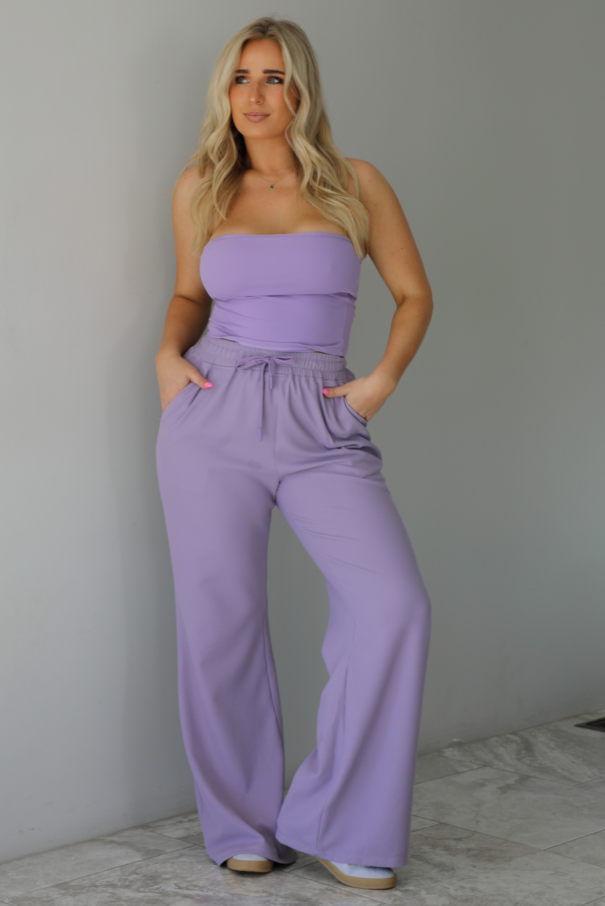 Woman wearing a two piece lavender set featuring a strapless, fitted tube top, and high waisted, flared hem pants. The photo is taken indoors against a white wall with white marble flooring.