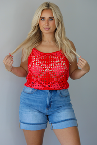 Woman wearing a pink and red loose knit crochet cropped top with light wash denim shorts indoors against a white wall.