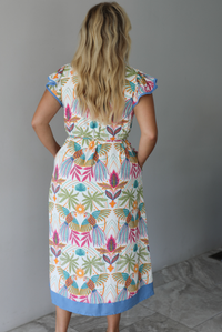 A woman wearing a short sleeved midi dress with a colorful tropical leaf print and light blue trim indoors against a white wall with white marble flooring. The photo shows the back of the dress. 