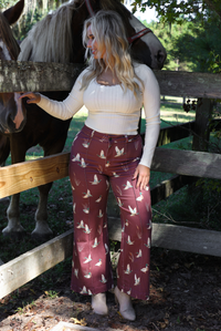 A woman wearing a cream top and maroon high waisted pants with a repeating mallard print throughout, standing outdoors against a wooden fence with horses and greenery in the background. 