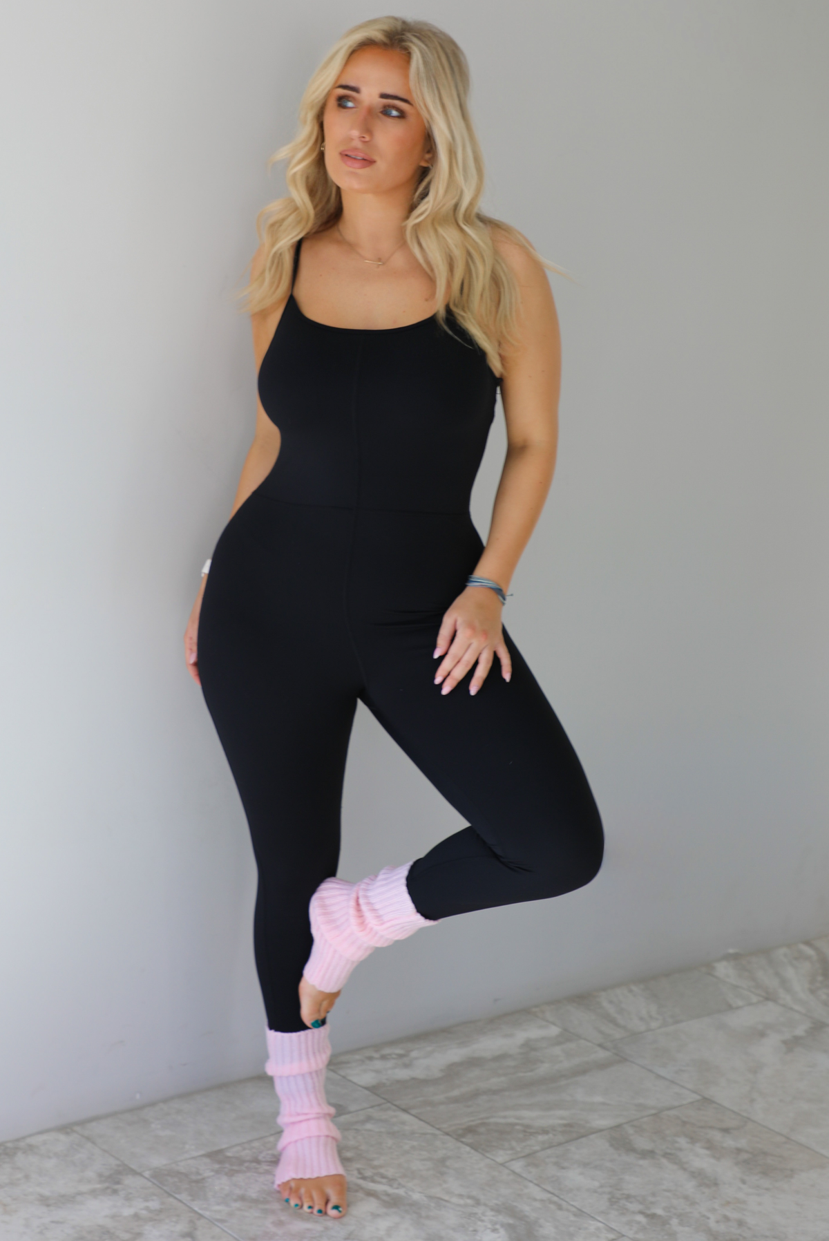 Woman wearing a black fitted jumpsuit with spaghetti straps. The jumpsuit is styled with pink leg warmers. The photo is taken indoors against a white wall with white marble flooring. 