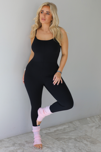 Woman wearing a black fitted jumpsuit with spaghetti straps. The jumpsuit is styled with pink leg warmers. The photo is taken indoors against a white wall with white marble flooring. 