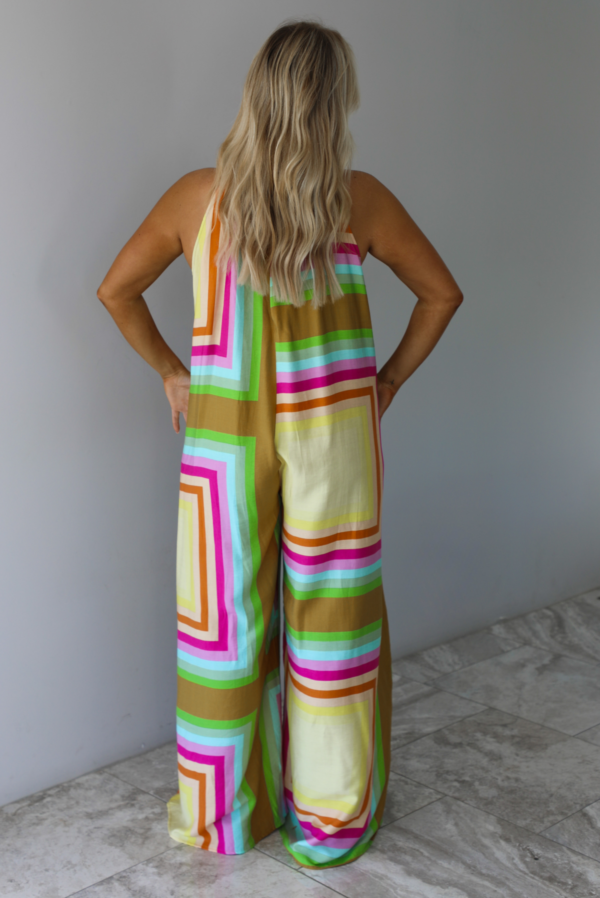 Woman wearing a colorful jumpsuit that has a high neckline, no sleeves, and a flowy cut with flared hem pants. The photo is taken indoors against a white wall with white marble flooring and shows the back of the jumpsuit. 
