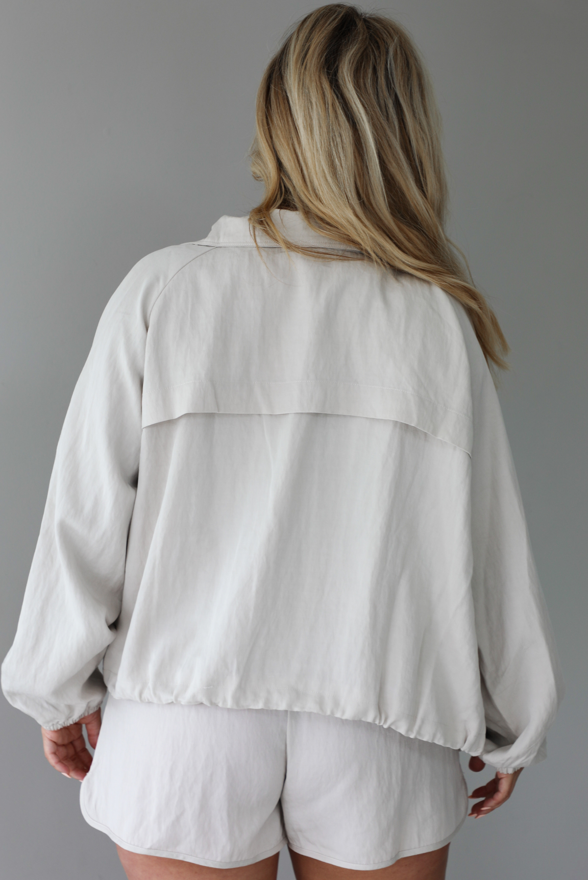 Woman wearing a stone colored two piece set featuring a zip up jacket and shorts with a navy tank tucked into the shorts indoors against a white wall. The photo shows the back of the set. 