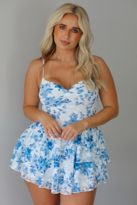 Woman wearing a white and blue floral mini dress with a low neckline, fitted bodice, and flowy, ruffled skirt indoors against a white wall.
