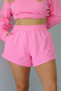 Woman wearing a hot pink two piece set featuring an oversized sweatshirt and high waisted shorts. The photo is taken indoors against a white wall and shows a closeup of the shorts of the set, which are high waisted, have an elastic waistline, and have functional pockets on either side. 