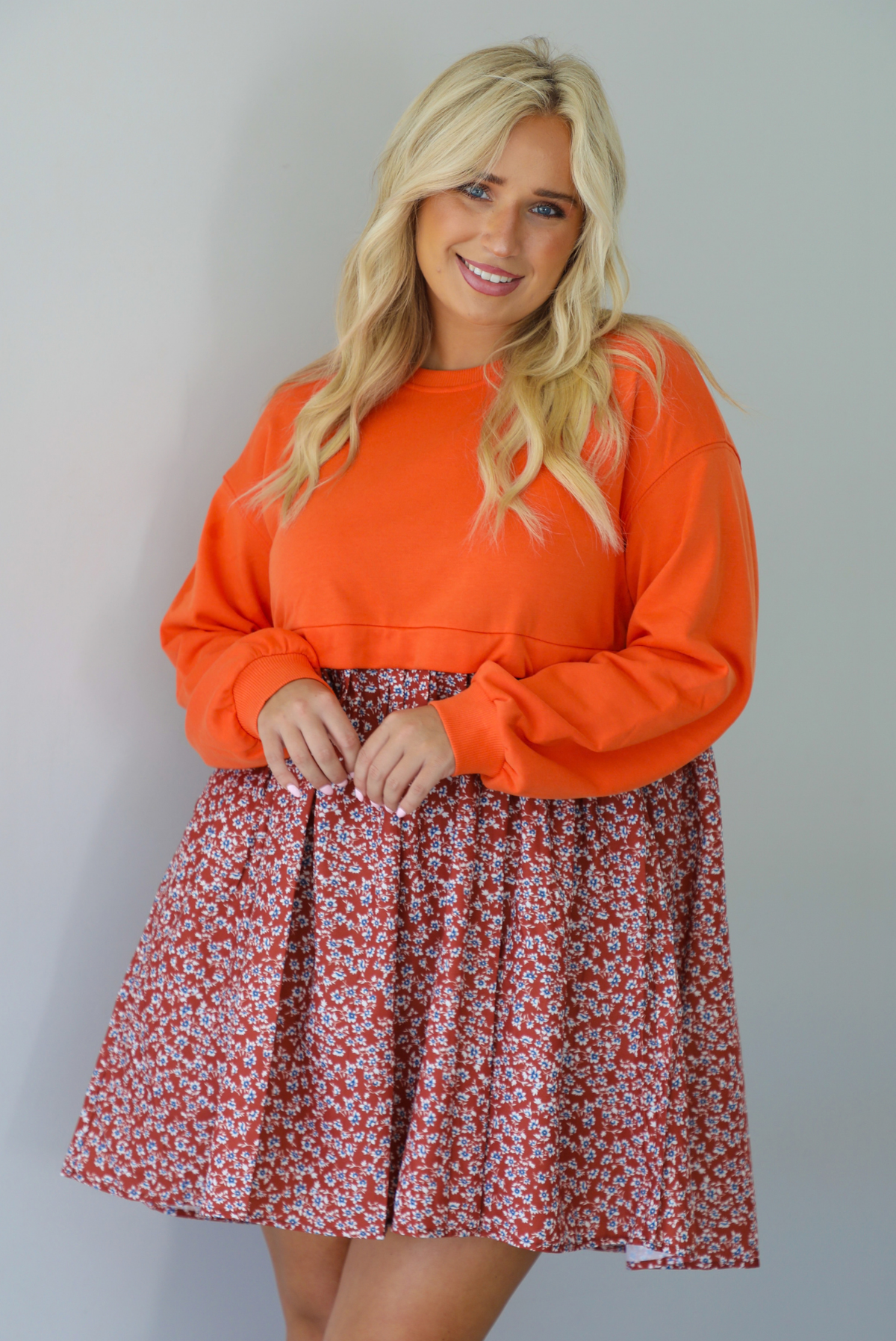 A woman wearing a block style oversized, flowy dress featuring a solid orange bodice and sleeves with a multi-colored floral skirt, standing indoors against a white wall.