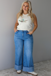A woman wearing a cream colored sweater vest with a mallard duck on the front, paired with medium wash denim with wide leg hem, standing indoors against a white wall with white marble flooring. 