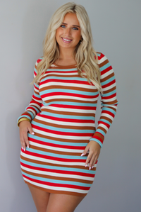 A woman wearing a red, blue, white, and brown striped long sleeved mini dress, standing indoors against a white wall.