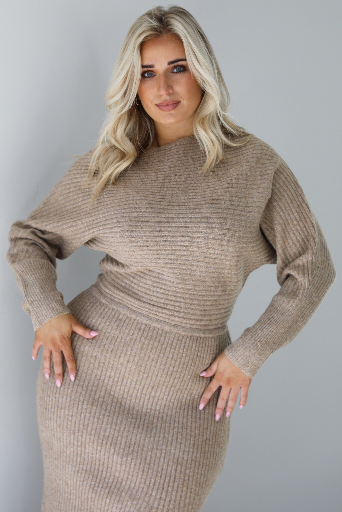 Woman wearing an oatmeal colored sweater dress that has a slouchy bodice that has long sleeves, with a fitted midi length skirt. The dress has a ribbed textured material that's horizontal on the bodice and vertical on the skirt. The photo is taken in front of a white wall.