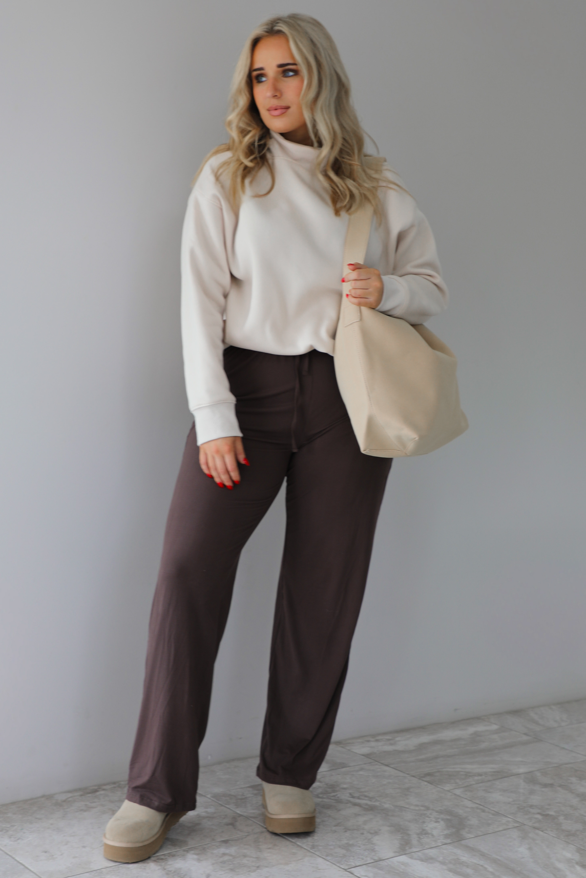 Woman wearing a beige high-neck, oversized sweater, high waisted brown pants, with a beige shoulder bag, and beige ugg boot shoes. The photo is taken indoors against a white wall with white marbled flooring. 