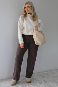 Woman wearing a beige high-neck, oversized sweater, high waisted brown pants, with a beige shoulder bag, and beige ugg boot shoes. The photo is taken indoors against a white wall with white marbled flooring. 