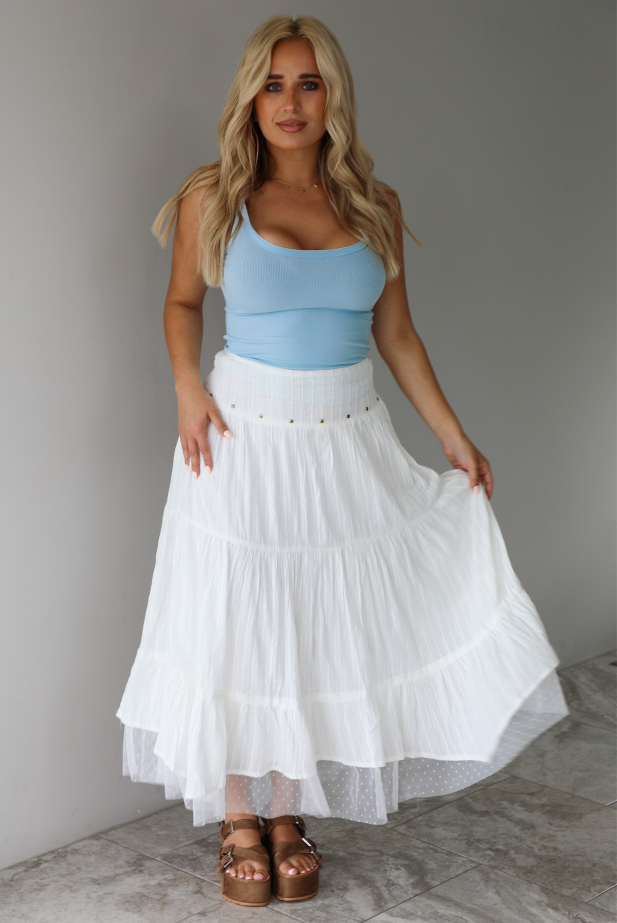 Woman wearing a blue tank and high waisted white maxi-length skirt indoors against a white wall with white marble flooring. 