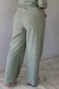 A woman wearing a pale green and brown striped two piece set featuring an oversized long sleeved top and high waisted, wide leg pants, standing indoors against a white wall with white marble flooring. The photo shows a closeup of the back of the pants, which have an elastic and drawstring waistline and functional pockets on either side.