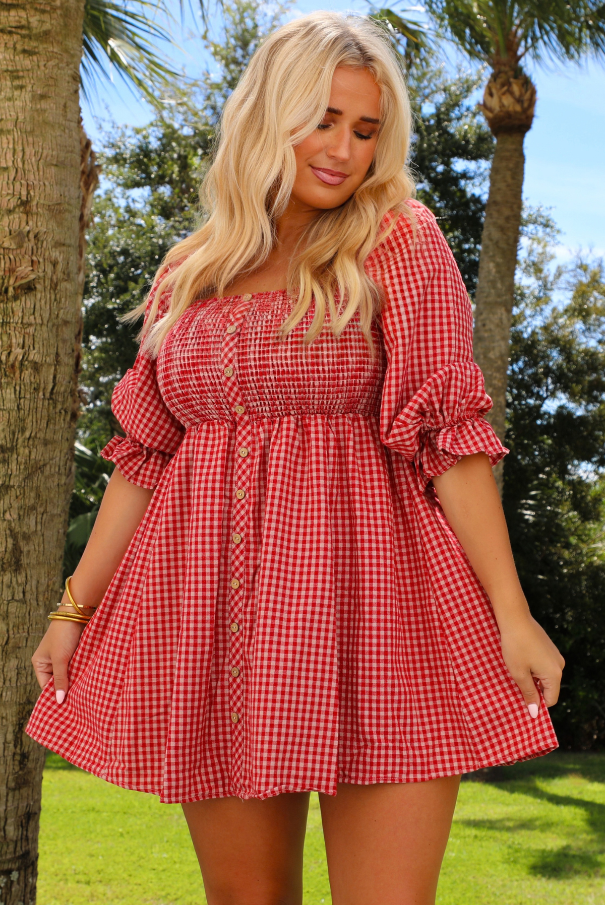 A woman wearing a red and white gingham mini dress with puff sleeves and a smocked bodice, standing outdoors in a grassy park with palm trees.
