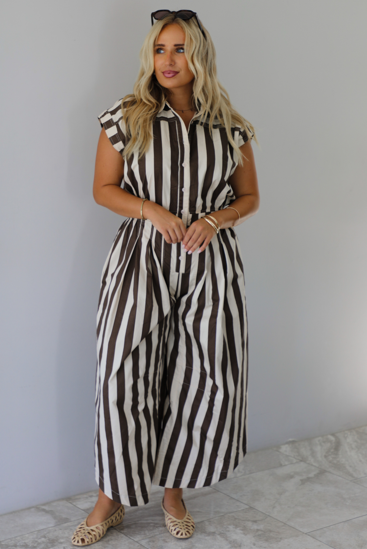 A woman wearing a brown and white striped patterned jumpsuit with a collared neckline, functional button closures in the front, a cinched waistline, and barrel-style bottoms, standing indoors against a white wall with white marble flooring. 