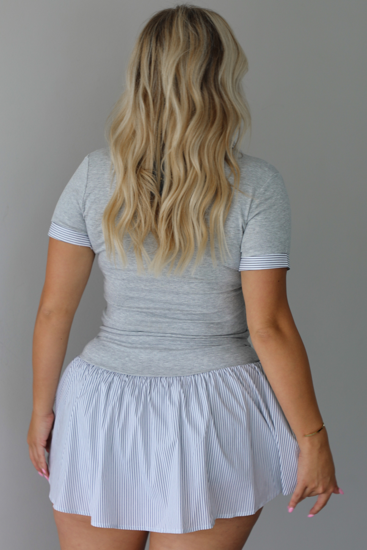 A woman shown from behind modeling a light gray short-sleeve collared top paired with a white flared mini skirt, styled with brown sandals, standing indoors against a white wall.