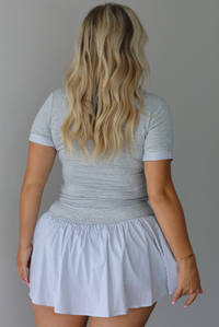 A woman shown from behind modeling a light gray short-sleeve collared top paired with a white flared mini skirt, styled with brown sandals, standing indoors against a white wall.