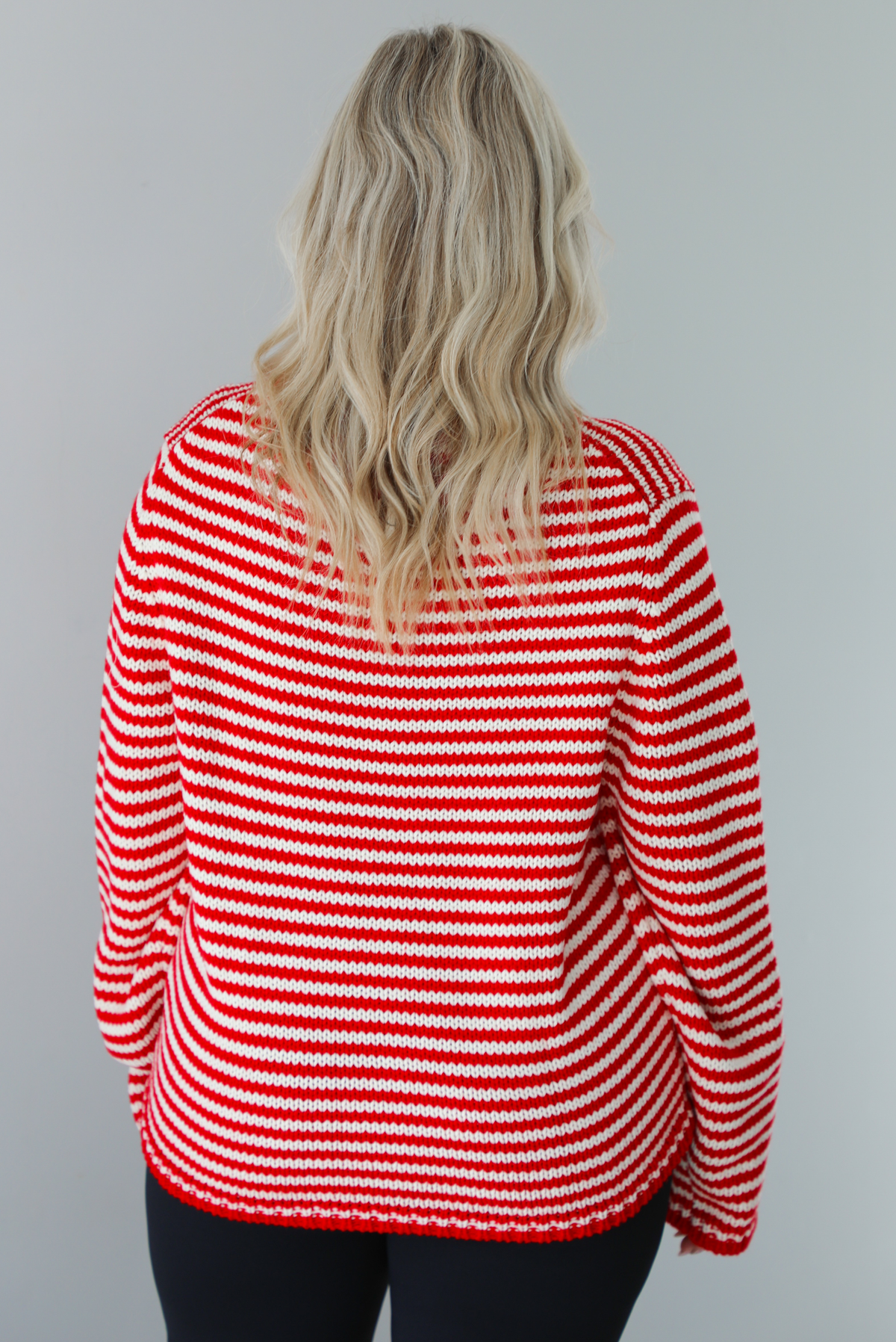 Woman wearing a white and red striped sweater that has a high neckline, an oversized fit, and is paired with black leggings. The photo shows a closeup of the back of the sweater. The photo is taken indoors against a white wall.